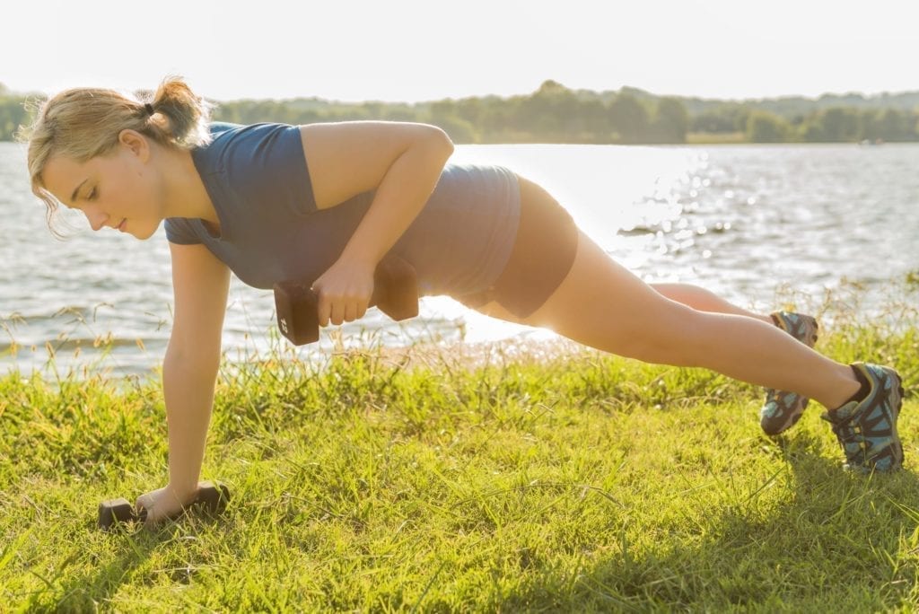 Young woman performing dumbbell row exercise lifting 12 pound weight in green grass outdoor park by river with soft sunlight